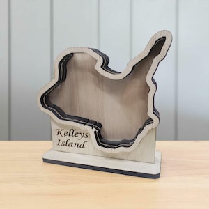 May include: A layered wooden map of Kelleys Island, with a light wood finish and dark edges. The island shape is cut out, creating a three-dimensional effect. The words "Kelleys Island" are engraved on the base.