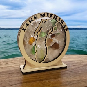 May include: A wooden cutout of the state of Michigan filled with sea glass, with the words "Lake Michigan" around the outside. The cutout is on a wooden base and is sitting on a wooden surface.