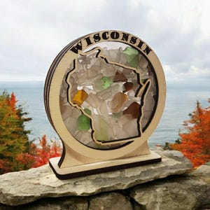 May include: A wooden cutout of the state of Wisconsin filled with colorful sea glass. The cutout is mounted on a wooden base and the words "WISCONSIN" are written around the top of the cutout.