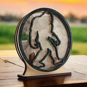 May include: A wooden Bigfoot silhouette with a clear resin insert filled with white and green crystals. The base is made of wood and has a dark blue stain.
