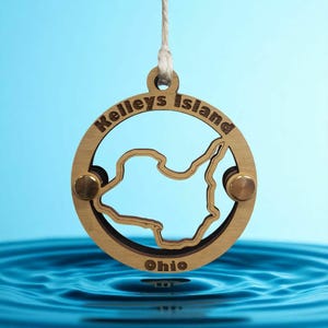 May include: A wooden ornament featuring the outline of Kelleys Island, Ohio, suspended against a blue background. The island shape is cut out within a circular frame, with the words "Kelleys Island" and "Ohio" engraved. The ornament is held by a string.
