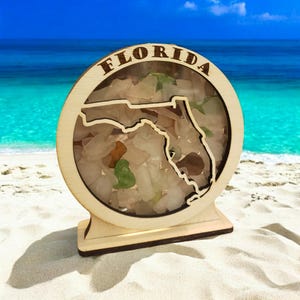 May include: A wooden display featuring the outline of Florida, filled with sea glass. The word "FLORIDA" is arched across the top. The display is set on a sandy beach with the ocean in the background.