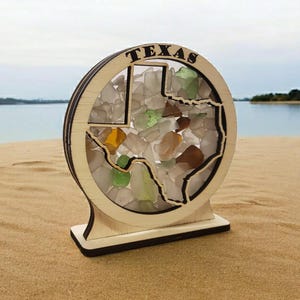 May include: A wooden cutout of the state of Texas filled with colorful sea glass. The cutout is mounted on a wooden base and has the word "TEXAS" written above the state.