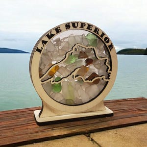 May include: A wooden cutout of Lake Superior with the words "Lake Superior" around the perimeter. The cutout is filled with small pieces of sea glass in various colors, including clear, green, and amber. The cutout is on a wooden base and is displayed against a background of a lake and sky.