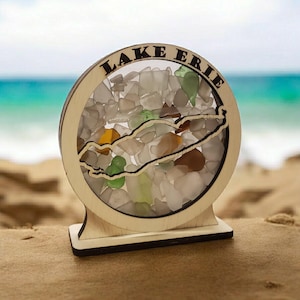May include: A wooden circular sign with the words "LAKE ERIE" inscribed around the edge. The sign features a cutout of the outline of Lake Erie filled with colorful sea glass.