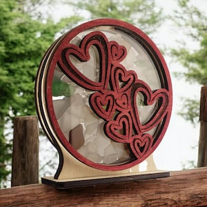 May include: A wooden heart-shaped ornament with a red finish and a clear glass center filled with small white and green stones. The hearts are layered and create a 3D effect.
