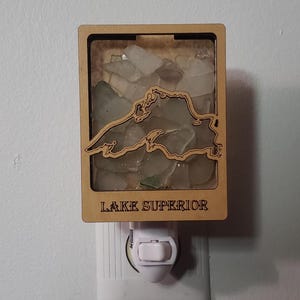 May include: A wooden night light shaped like Lake Superior, filled with clear and green sea glass. The night light has a switch on the bottom and says "LAKE SUPERIOR" on the front.