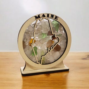 May include: A wooden cutout of the state of Maine filled with sea glass. The cutout is mounted on a wooden base and has the word "MAINE" written above the state.