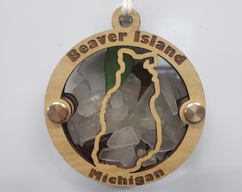 Puede incluir: Un adorno redondo de madera con las palabras "Beaver Island Michigan" grabadas en el borde. El centro presenta un recorte de la forma de la isla, lleno de vidrio de mar transparente y verde. Dos remaches color latón aseguran el marco de madera.