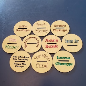 May include: Nine wooden round coin banks with different labels, including "Life Savings", "Beach Money", "Spare Change", "Fun Money", "Retirement Fund", "Ava's Bank", "Swear Jar", "She who does the laundry keeps the change", and "Vacation Fund".