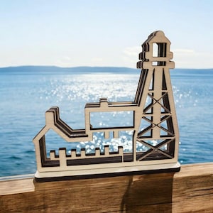 May include: A wooden cutout of a lighthouse with a building next to it. The cutout is brown and has a white background. The lighthouse has a tall tower with a small roof and a window. The building has a rectangular shape with a small roof and a window. The cutout is sitting on a wooden surface.