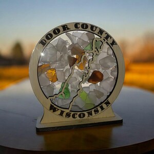 May include: A wooden Door County, Wisconsin, sign with a circular design. The sign features the outline of Door County filled with sea glass in various colors. The words "Door County" and "Wisconsin" are etched around the circle.