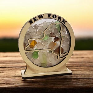May include: A wooden cutout of the state of New York filled with colorful sea glass. The cutout is mounted on a wooden base and has the words "NEW YORK" written around the edge.