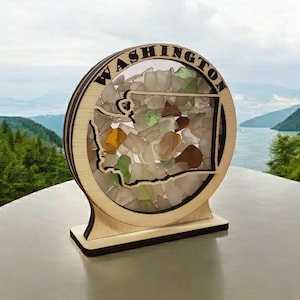 May include: Wooden cutout of the state of Washington filled with colorful sea glass. The cutout is mounted on a wooden base and has the word "WASHINGTON" written around the edge.