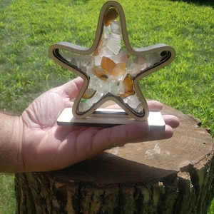 May include: A wooden starfish-shaped frame filled with sea glass in shades of green, white, and orange. The frame is on a wooden stand.