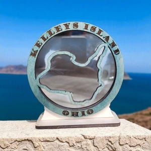 May include: A wooden souvenir featuring a teal-colored frame with the words "KELLEY'S ISLAND OHIO" encircling a clear window. The window showcases the island's outline. The base is white and brown, set against a scenic backdrop of water and sky.