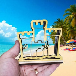 May include: Wooden sandcastle-shaped decoration with a beach scene visible through the cutouts. The sandcastle is made of light-colored wood and features a seashell and starfish design. The background shows a blue sky, ocean, and palm trees.