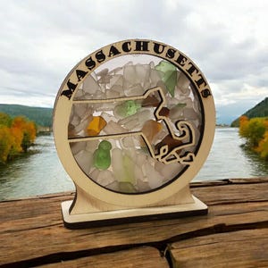 May include: A wooden cutout of the state of Massachusetts filled with colorful sea glass. The cutout is surrounded by a circle with the word "MASSACHUSETTS" written around it. The cutout is on a wooden stand.