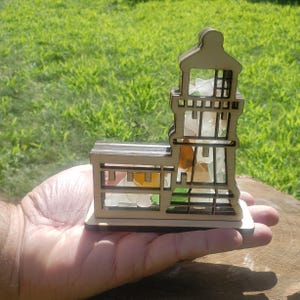 May include: A wooden lighthouse model with a glass mosaic design. The lighthouse is made of light brown wood and has a clear glass window. The glass mosaic is made of small pieces of colored glass in shades of green, blue, and white.