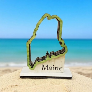 May include: A wooden decorative piece in the shape of the state of Maine, painted green and brown, with the word "Maine" at the base. The piece is set on a white base, with a beach and ocean background.