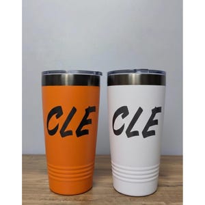 Puede incluir: Dos vasos aislados, uno naranja y otro blanco, cada uno con un gráfico "CLE" negro. Los vasos tienen un borde de acero inoxidable y una tapa negra. Los vasos son cilíndricos y miden unos 18 cm de alto.