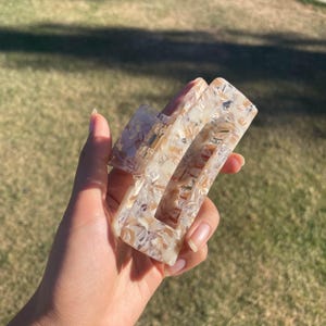 May include: A rectangular hair claw clip in a cream and tan marbled pattern. The clip has a spring mechanism and is held in a hand against a blurred green background. The clip is approximately 4 inches long.
