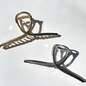 May include: Two metal hair claw clips. One is gold-toned, the other is silver-toned. Both have a curved design with a spring mechanism and small teeth along the edges for grip. The clips are on a white surface.