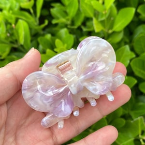 May include: A large, iridescent purple and white plastic hair claw with a bow design. The claw has a gold-toned metal clasp.
