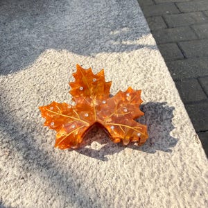 Peut inclure: Une pince à cheveux en forme de feuille d'érable orange avec des strass incrustés. La pince est faite d'un matériau translucide et présente des veines dorées. Elle est conçue pour maintenir les cheveux en place.