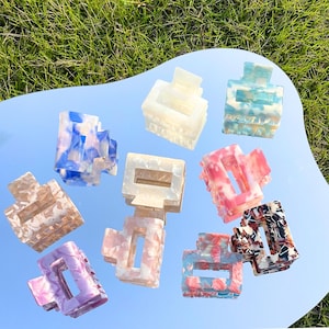 Könnte beinhalten: Eine Sammlung von zehn rechteckigen Haarspangen mit verschiedenen Marmormustern in Farben wie Blau, Weiß, Rosa, Lila, Beige und Schwarz. Jede Spange verfügt über ein Klappdesign mit ineinandergreifenden Zähnen zum Befestigen der Haare.