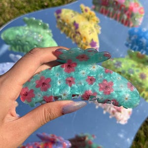 Pode incluir: Uma presilha de cabelo turquesa com um padrão floral de flores rosa e margaridas brancas. A presilha tem uma borda ondulada. Outras presilhas de cabelo coloridas são visíveis no fundo.