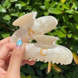 May include: Two pearlescent hair claw clips with gold letter accents. One clip has the letter "A" and the other has the letter "C". The clips are a light beige color with a marbled effect.