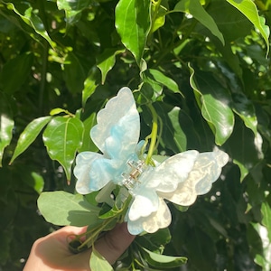 Puede incluir: Una pinza para el pelo de color azul claro en forma de mariposa con un acabado brillante. La pinza está hecha de plástico y tiene un cierre de metal.