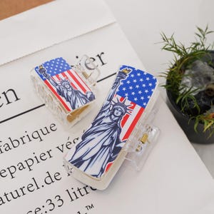 May include: Two clear plastic hair clips with a design of the Statue of Liberty and the American flag. The Statue of Liberty is depicted in blue and white, while the American flag is red, white, and blue.