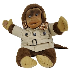 May include: A soft toy monkey with brown fur, dressed in a tan jacket with a black belt and buttons. The monkey has a brown face and a leopard print scarf. Its arms are outstretched, and it has tan hands and feet.