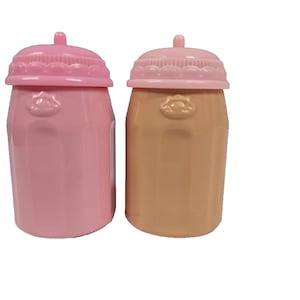 May include: Two baby bottles, one pink and one tan, with matching pink lids. Each bottle has a small, rounded top and a decorative design on the front. The lids have a small, rounded nipple-like feature.