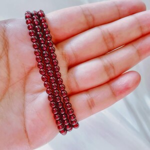 May include: A strand of dark red garnet beads, strung on a clear thread, is displayed on a hand. The beads are small and round, and they have a smooth, polished finish.