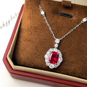 May include: A silver necklace with a large, square, red gemstone pendant surrounded by smaller, clear gemstones. The necklace is displayed in a red jewelry box with a brown interior.