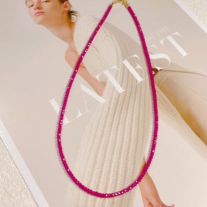 May include: A vibrant magenta beaded necklace with a gold-colored clasp. The necklace is draped against a cream-colored sweater and a magazine with the word "LATEST" printed on it.