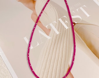 May include: A vibrant magenta beaded necklace with a gold-colored clasp. The necklace is draped against a cream-colored sweater and a magazine with the word "LATEST" printed on it.