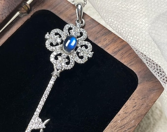 May include: A silver key pendant necklace with a blue gemstone center. The key-shaped pendant is encrusted with small, clear crystals and hangs from a delicate silver chain. The necklace is displayed in a wooden jewelry box with a black velvet interior.