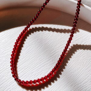 May include: A red gemstone necklace made of small, round beads. The necklace is lying on a white surface.