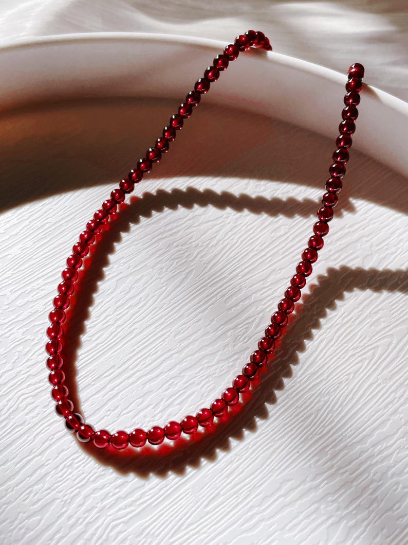 May include: A red gemstone necklace made of small, round beads. The necklace is lying on a white surface.