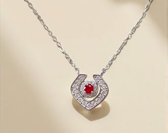 May include: A silver necklace with a horseshoe-shaped pendant. The pendant features a central red gemstone surrounded by a halo of smaller, clear gemstones. The chain is delicate and silver.