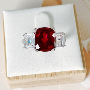 May include: A silver ring featuring a large, cushion-cut, deep red gemstone flanked by two rectangular, clear gemstones. The ring is set on a white display stand.