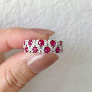 May include: A silver ring with two rows of round, vibrant red gemstones, interspersed with smaller, clear stones. The ring is held against a soft, neutral background, highlighting the sparkle of the gems.