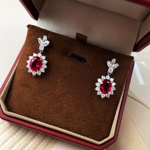 May include: A pair of elegant drop earrings displayed in a red jewelry box. Each earring features a large, oval-shaped red gemstone surrounded by a halo of smaller, clear stones. A cluster of clear stones tops each earring.