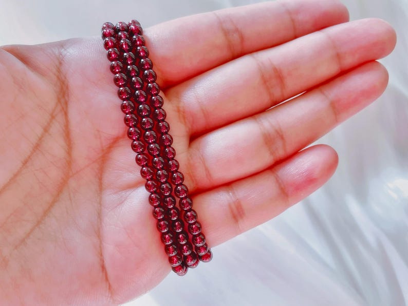 May include: A strand of dark red garnet beads, strung on a clear thread, is displayed on a hand.