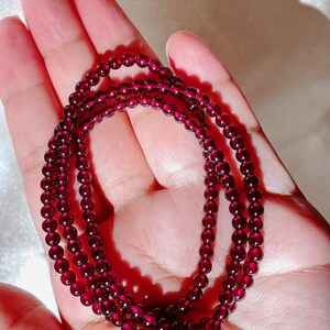 May include: A strand of dark red garnet beads, strung on a clear thread, is displayed on a hand. The beads are small and round, and the strand is long enough to wrap around the wrist multiple times.