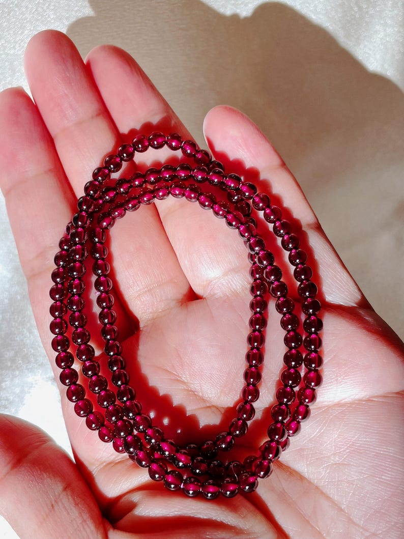 May include: A strand of dark red garnet beads, strung on a clear thread, is displayed on a hand. The beads are small and round, and the strand is long enough to wrap around the wrist multiple times.